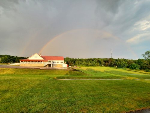 A white building with a red roof under a cloudy sky, featuring a rainbow arching over green fields.