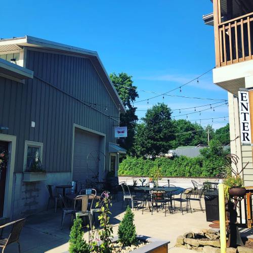 A sunny outdoor patio with tables and chairs, string lights overhead, surrounded by greenery and buildings.