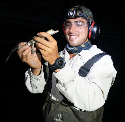 A person wearing a headlamp and protective gear holds a small crocodile, smiling in a dark environment.
