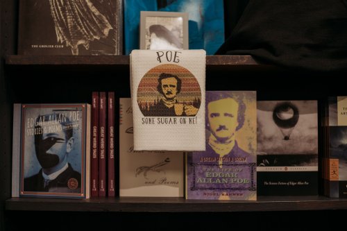 A shelf displaying books about Edgar Allan Poe, with a towel featuring his image and the text "Poe: Some Sugar on Me."