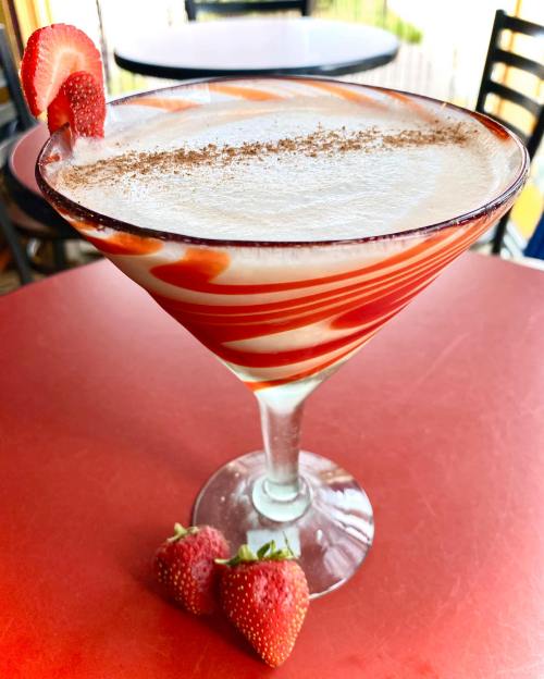 A colorful cocktail in a glass, garnished with a strawberry and drizzled with syrup, sits on a red table.