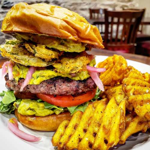 A towering burger with avocado, pickled onions, and crispy toppings, served with a side of waffle fries.