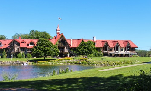 A scenic view of a large lodge with a red roof, surrounded by greenery and a calm lake under a clear blue sky.