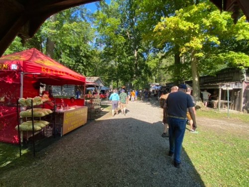 A bustling outdoor market with colorful tents, vendors, and visitors walking along a gravel path surrounded by trees.