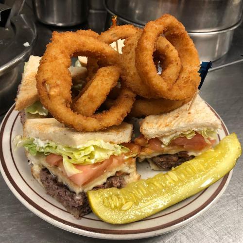 A stacked sandwich with lettuce, tomato, and bacon, topped with onion rings and served with a pickle on a plate.