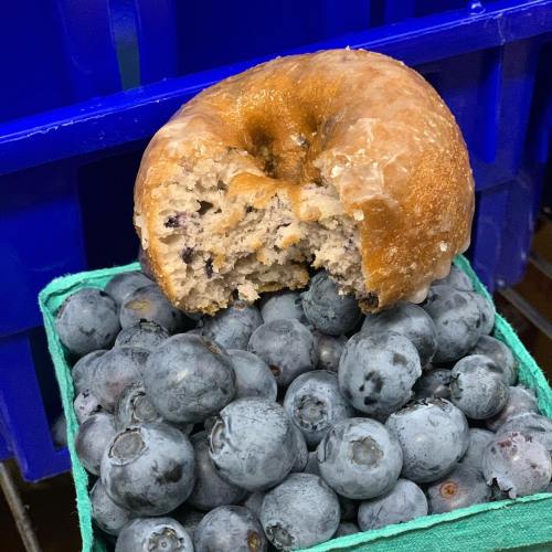 A glazed donut with a bite taken out, resting on a bed of fresh blueberries in a green basket.