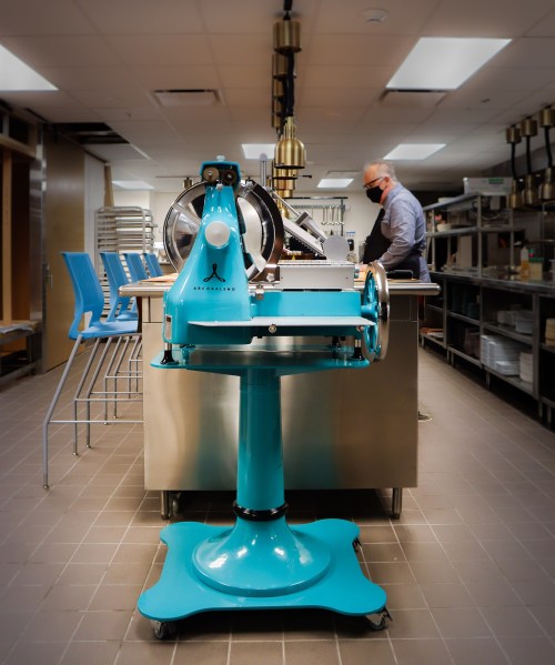 A turquoise kitchen machine on wheels in a modern kitchen with a person wearing a mask in the background.