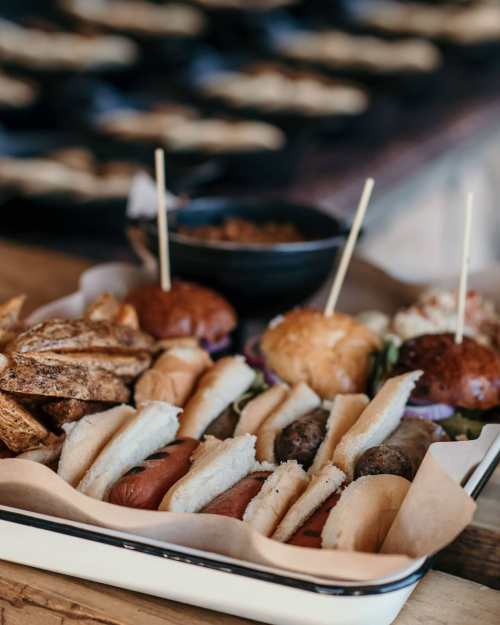 A tray of assorted hot dogs and sliders, accompanied by crispy potato wedges and a small bowl of sauce.