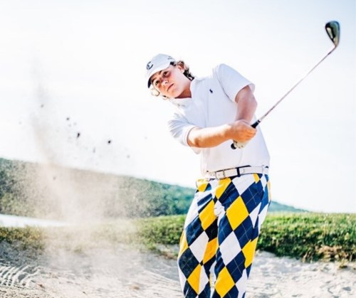 A young golfer swings a club in a sand trap, wearing a white shirt and colorful checkered pants.