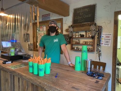 A staff member in a green shirt and mask stands behind a wooden counter at Indian Creek Zoo, with colorful cups and snacks.