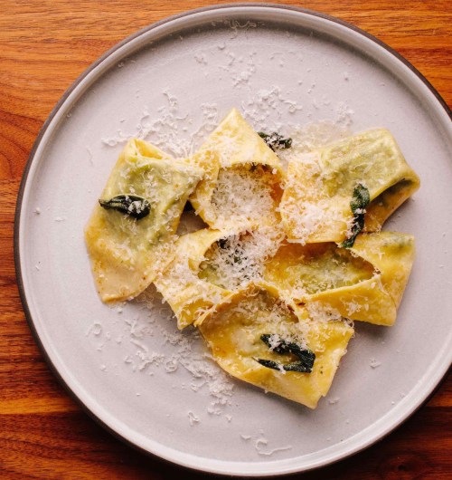 A plate of ravioli topped with grated cheese and fresh sage on a wooden surface.