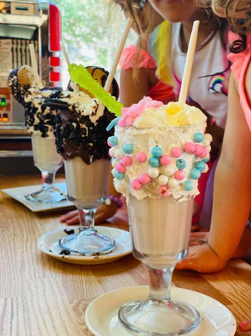Three colorful milkshakes topped with whipped cream, candies, and cookies, with a child in the background.