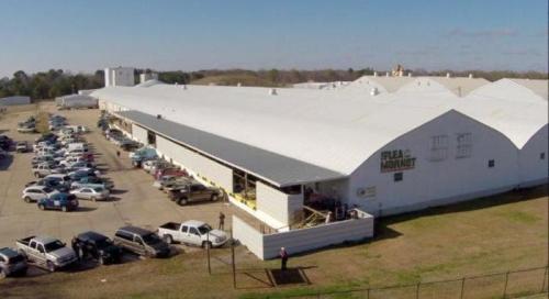 Aerial view of a large flea market building with parked cars outside on a sunny day.
