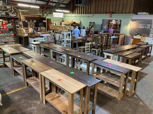 A spacious workshop filled with wooden tables and chairs, with colorful price tags on each piece. People browse in the background.