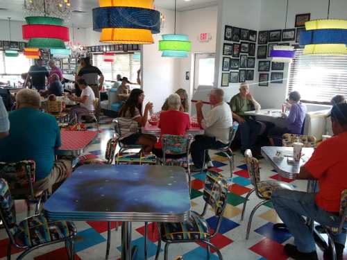 A colorful diner filled with people dining and chatting, featuring vibrant decor and a retro atmosphere.