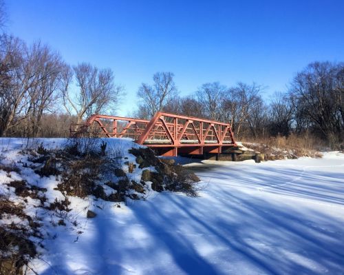 A red metal bridge spans a frozen river, surrounded by snow and bare trees under a clear blue sky.