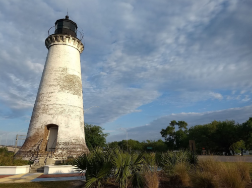 A tall, weathered lighthouse stands against a cloudy sky, surrounded by greenery and palm plants.