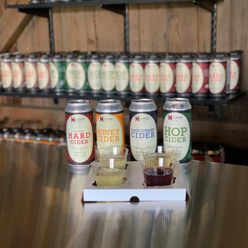 A selection of cider cans on a shelf with four tasting cups in front, showcasing different cider flavors.