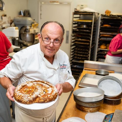A chef holds a large pastry in a bakery, with baking equipment and staff visible in the background.