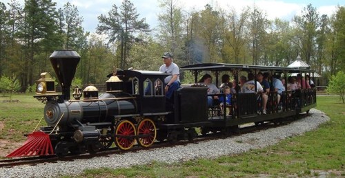 A vintage steam train pulls a passenger car along a gravel track through a green landscape with trees.