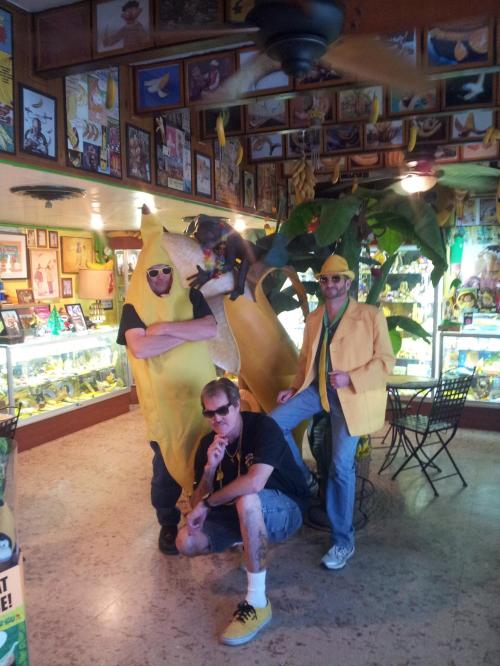 Three people in banana-themed costumes pose in a colorful room decorated with banana art and plants.
