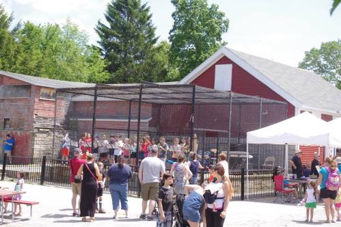 A crowd gathers near a fenced area with children performing, surrounded by trees and a red barn in the background.
