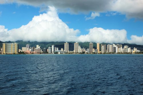 A panoramic view of a city skyline along the coast, with tall buildings and mountains under a partly cloudy sky.