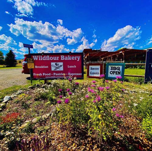 Sign for Wildflour Bakery with flowers in front, indicating breakfast, lunch, and BBQ options; bakery closed. Bright blue sky.