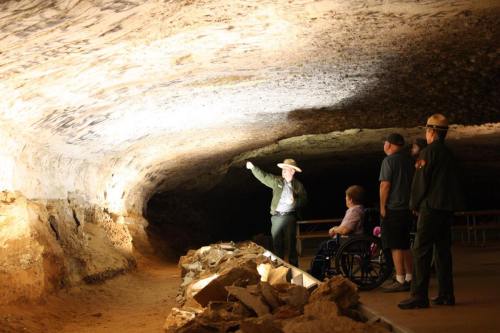 A park ranger points out features in a cave while visitors, including a person in a wheelchair, listen attentively.