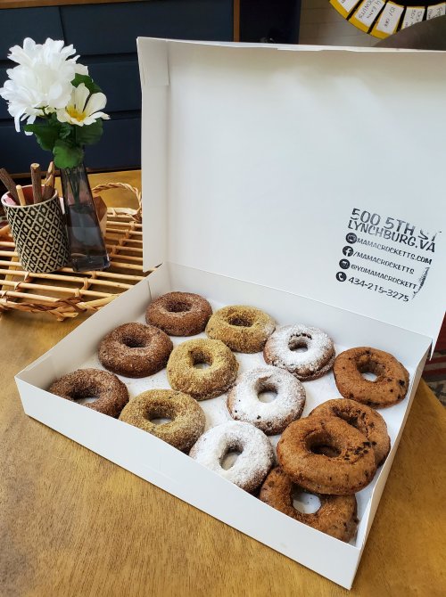 A box of assorted donuts on a wooden table, with a flower vase and utensils in the background.