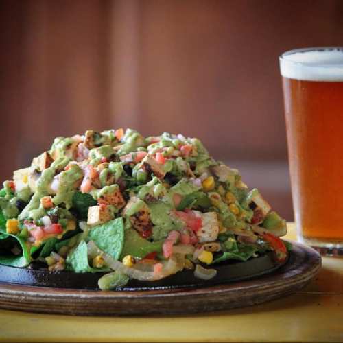 A vibrant salad with grilled chicken, avocado dressing, and colorful toppings, served with a glass of beer.