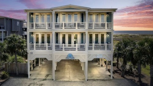A beachfront house with multiple balconies, surrounded by palm trees, under a colorful sunset sky.
