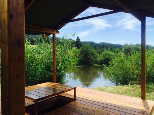 A wooden deck with a bench overlooks a serene river surrounded by lush greenery and distant hills under a blue sky.