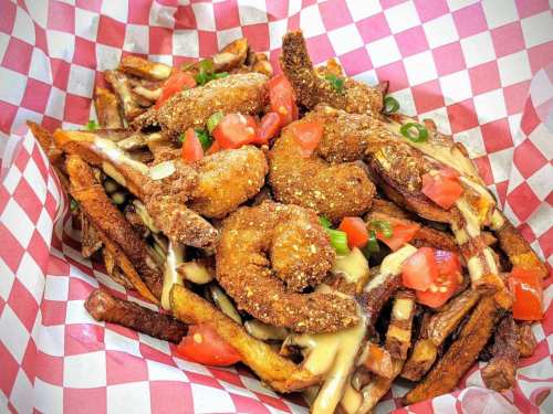 A basket of crispy fries topped with fried shrimp, diced tomatoes, and drizzled with sauce, all in a checkered paper.