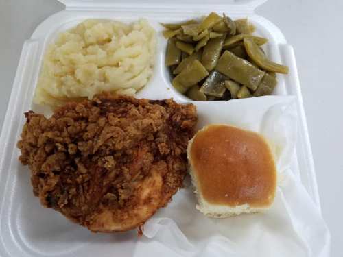 A takeout container with fried chicken, mashed potatoes, green beans, and a dinner roll.