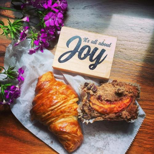 A croissant and muffin on parchment paper, with purple flowers and a sign that says "It's all about Joy."