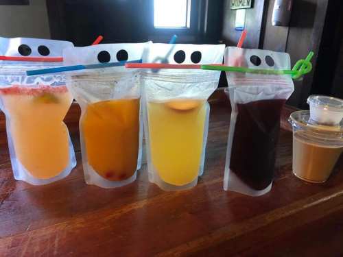 Four colorful drinks in pouches with smiling faces, lined up on a wooden surface, alongside two small containers.