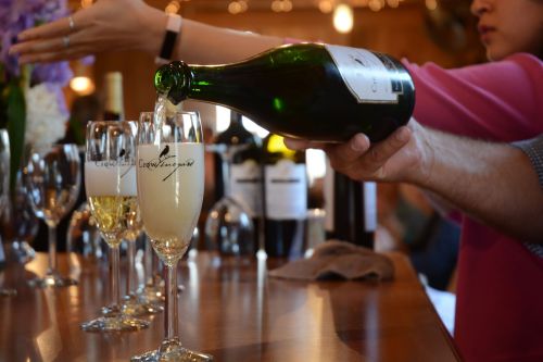 A hand pours sparkling wine into a flute at a bar, with several glasses and bottles in the background.