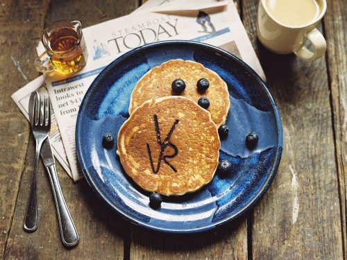 Two pancakes with blueberries and chocolate drizzle on a blue plate, accompanied by a newspaper and a cup of coffee.
