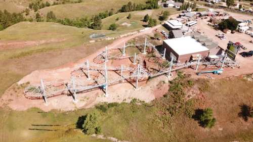 Aerial view of a roller coaster on a hillside, with a building nearby and green landscape in the background.