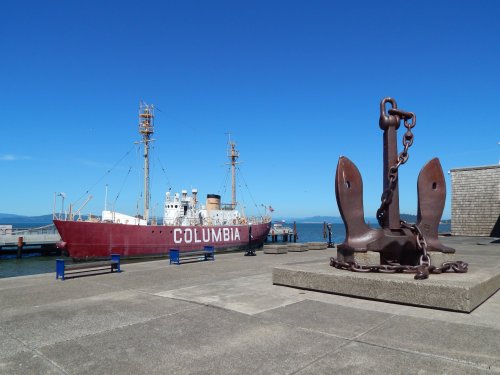 A red ship named "Columbia" docked at a pier, with a large anchor in the foreground against a clear blue sky.