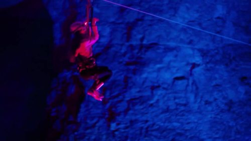 A person rappelling down a rock face illuminated by blue and purple lights.