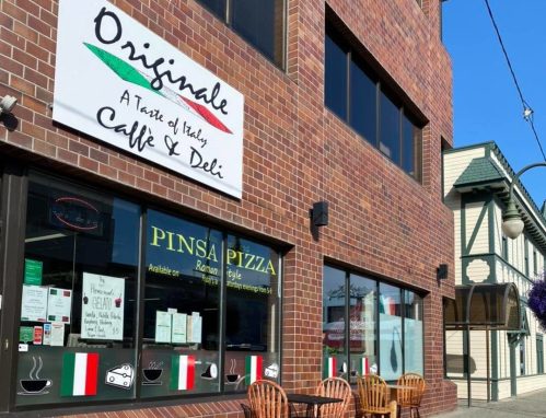 Exterior of a brick building featuring a café and deli sign, with outdoor seating and a "Pinsa Pizza" window display.