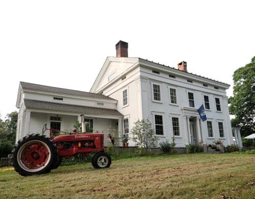 A classic white farmhouse with a red tractor in the foreground, surrounded by a grassy yard and trees.