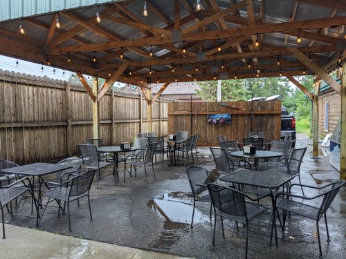 Outdoor seating area with empty tables under a wooden gazebo, surrounded by a wooden fence and wet pavement.