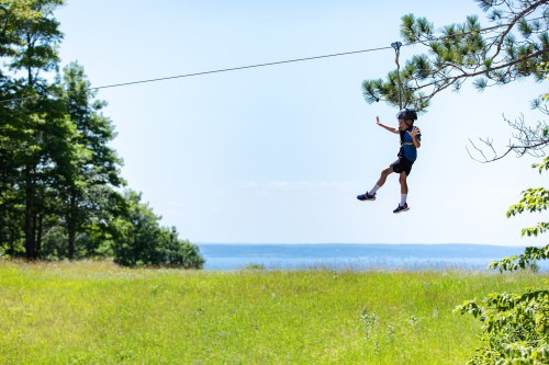 A child on a zip line glides over a grassy field with trees and a lake in the background under a clear blue sky.