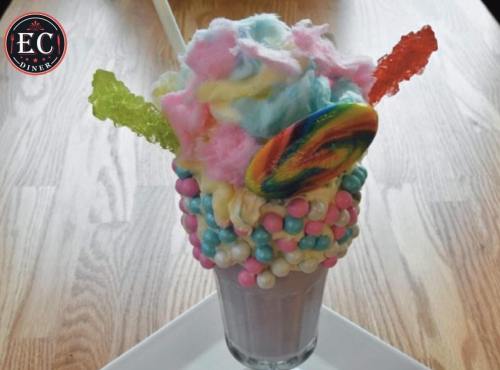 A colorful milkshake topped with cotton candy, a lollipop, and candy sticks, served in a glass on a wooden table.