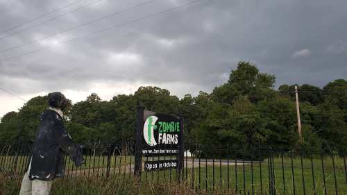 A spooky scarecrow stands near a sign for Zombie Farms under dark, cloudy skies.