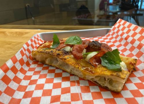 A slice of pizza on a red and white checkered paper, topped with sauce, cheese, and fresh basil.