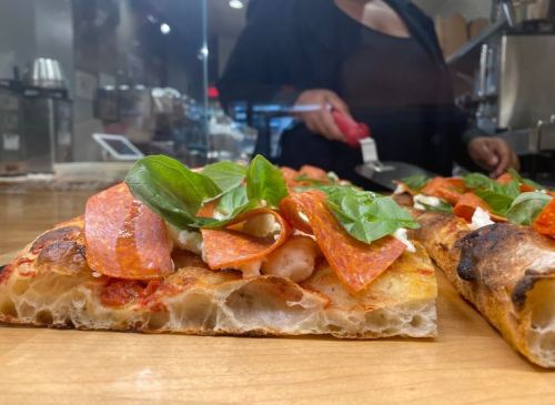 A close-up of a slice of pizza topped with pepperoni and fresh basil, with a person preparing food in the background.
